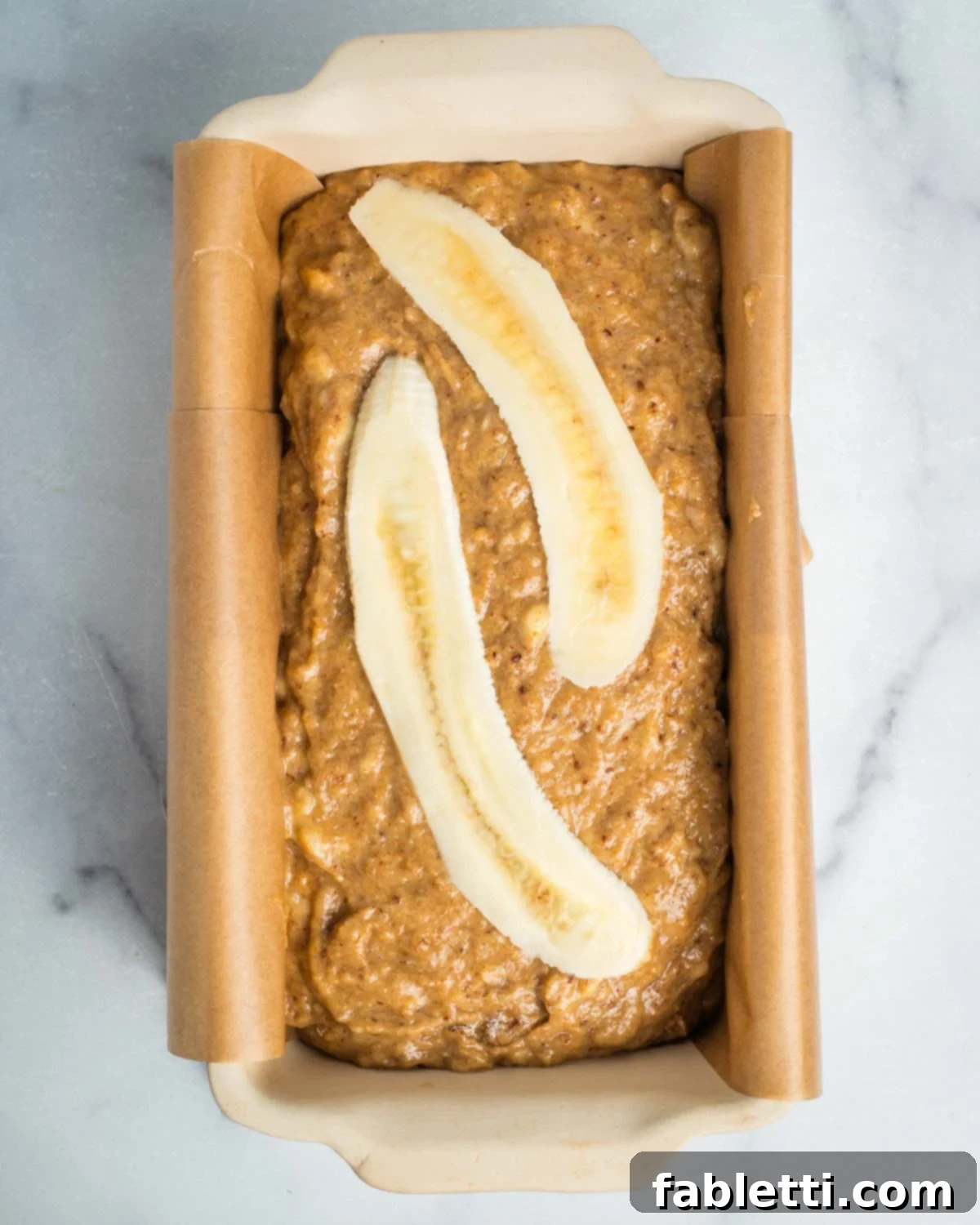 Banana bread batter in a loaf pan, decorated aesthetically with two long, thin slices of fresh banana placed on top before baking.