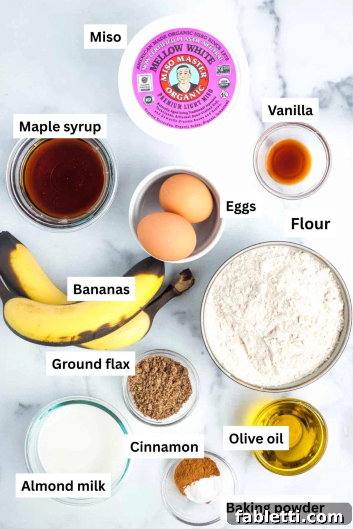 Assortment of key ingredients for banana bread including miso, bananas, eggs, flour, and maple syrup.