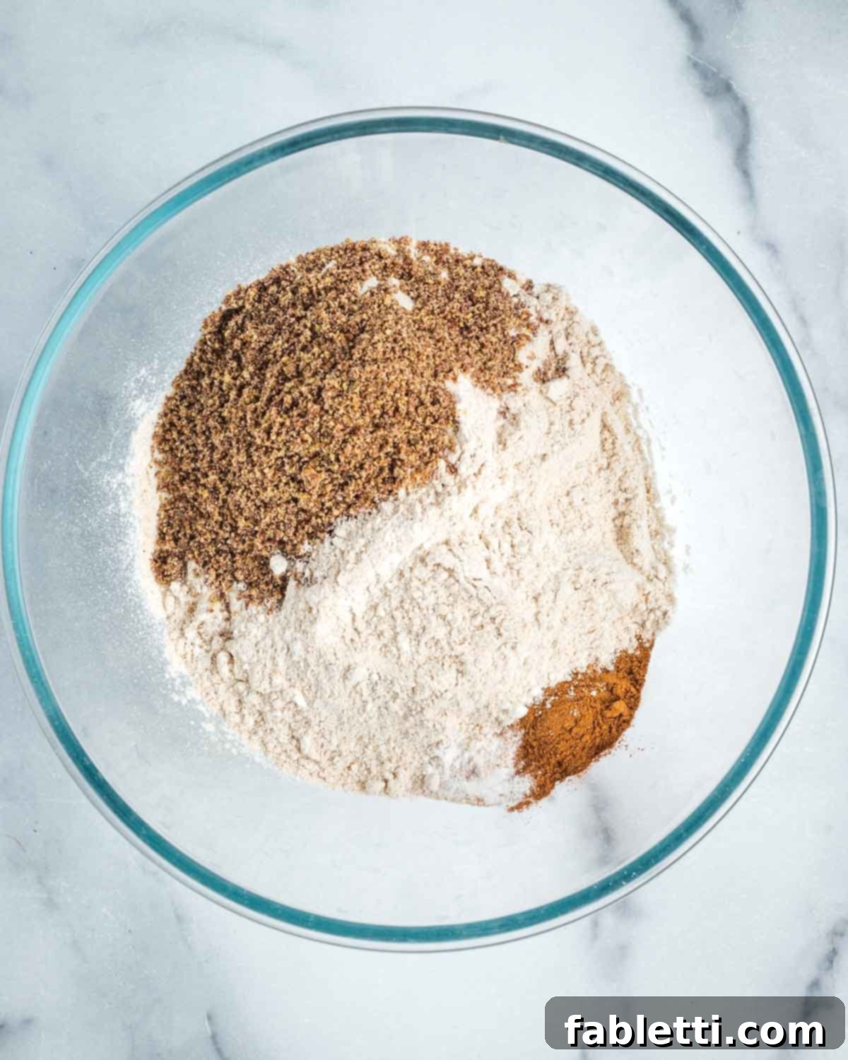 Flour, ground flax, cinnamon, and baking powder are measured into a clear glass mixing bowl, ready to be combined for the banana bread.