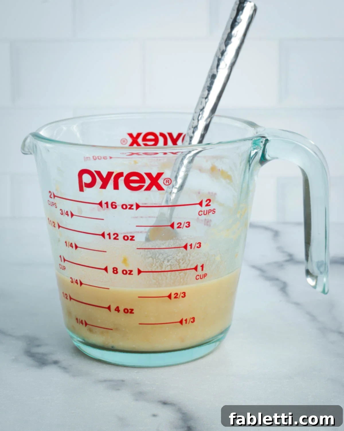 A fork is used to mash and disperse miso paste into almond milk along the sides of a glass measuring cup.