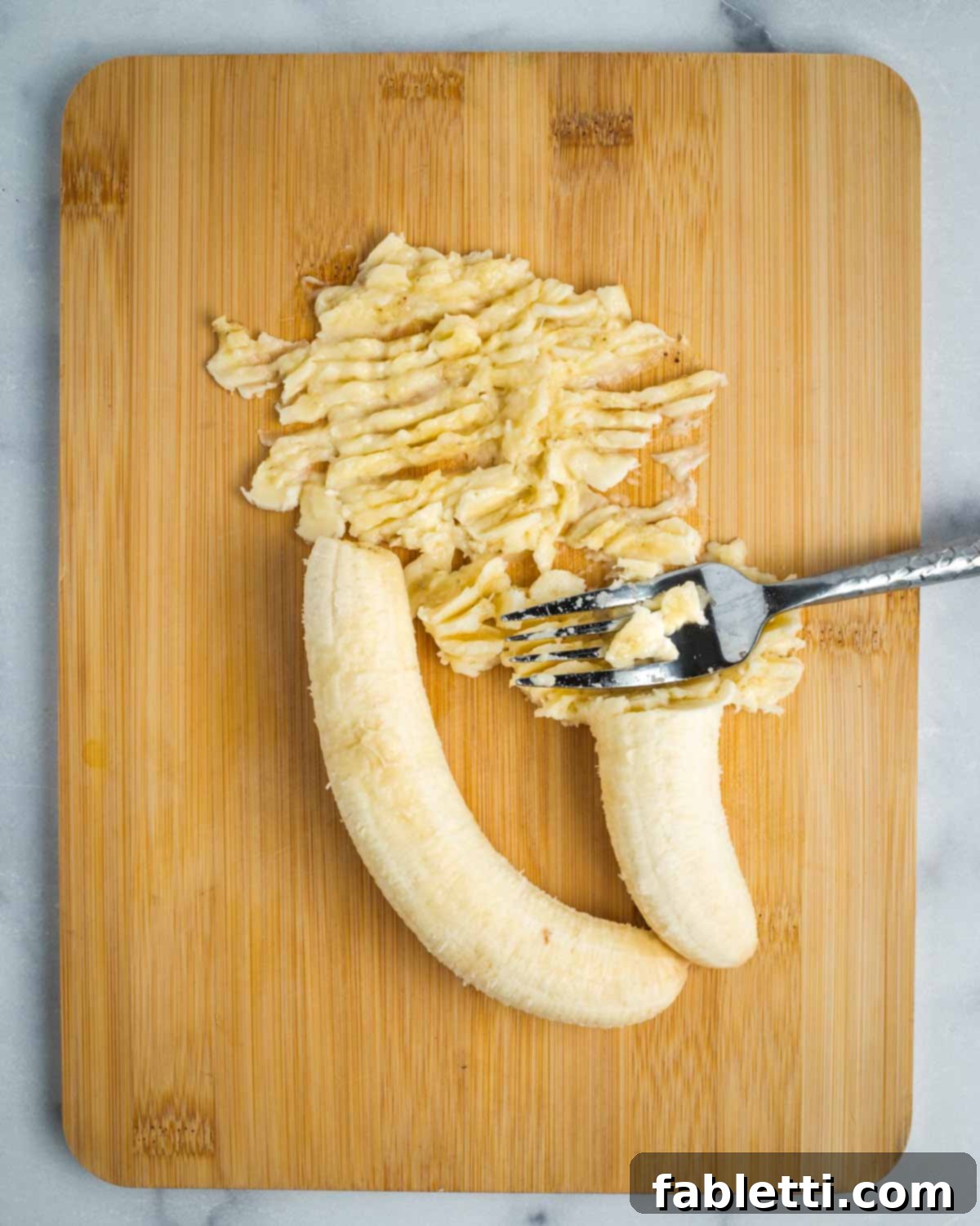 Ripe bananas being mashed with a fork on a wooden cutting board, illustrating the initial mashing process.