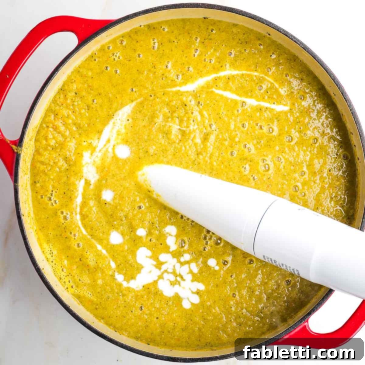 Velvety Plant-Based Broccoli Soup 12 Immersion blender pureeing soup with a streak of white cream throughout.