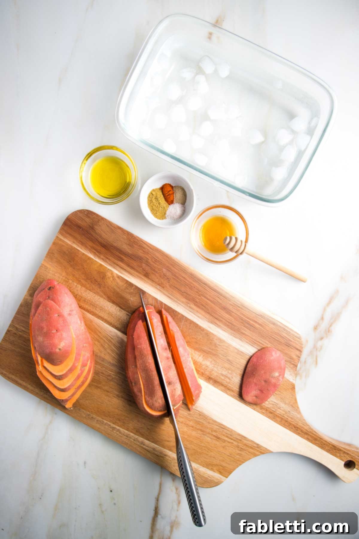 Golden Crispy Sweet Potato Fries 5 Cutting sweet potatoes into fries on a cutting board. First sliced one way, then the other. Along with glass bowls with olive oil, spices and honey, plus an ice water bath waiting to soak the fries.