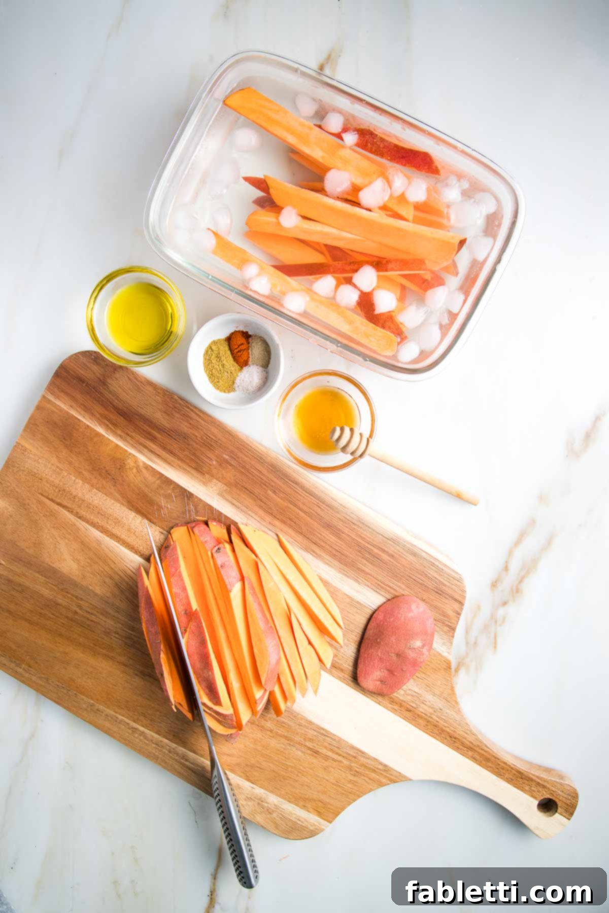 Golden Crispy Sweet Potato Fries 6 Sweet potato cut into fries on a cutting board. Some are in a large dish of ice water. Olive oil, spices and honey in small glass dishes.