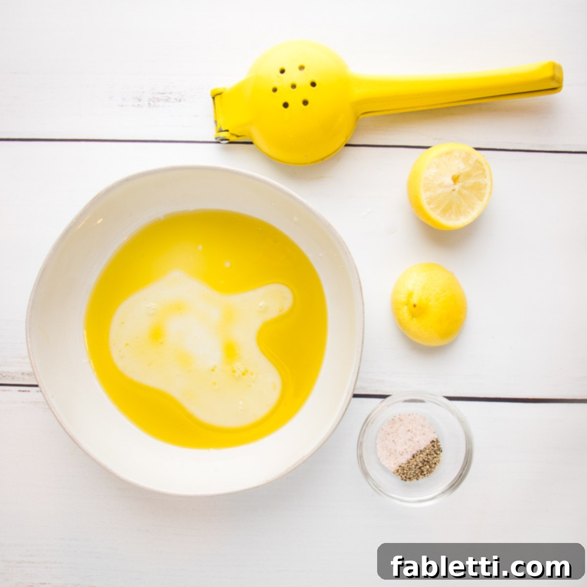 Vibrant Arugula & Lemon Salad 6 A white bowl holds separated olive oil and freshly squeezed lemon juice, ready to be emulsified. Beside it, two lemon halves and a yellow citrus juicer, along with a small dish containing salt and pepper, complete the setup.