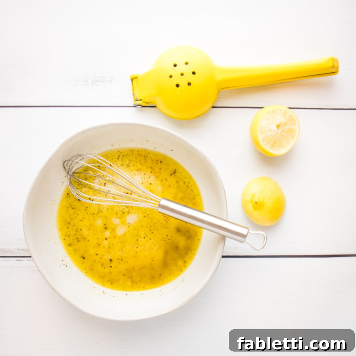 Vibrant Arugula & Lemon Salad 7 A lemon vinaigrette is being expertly emulsified in a white bowl using a whisk, with two squeezed lemon halves visible on the side, indicating the fresh preparation.