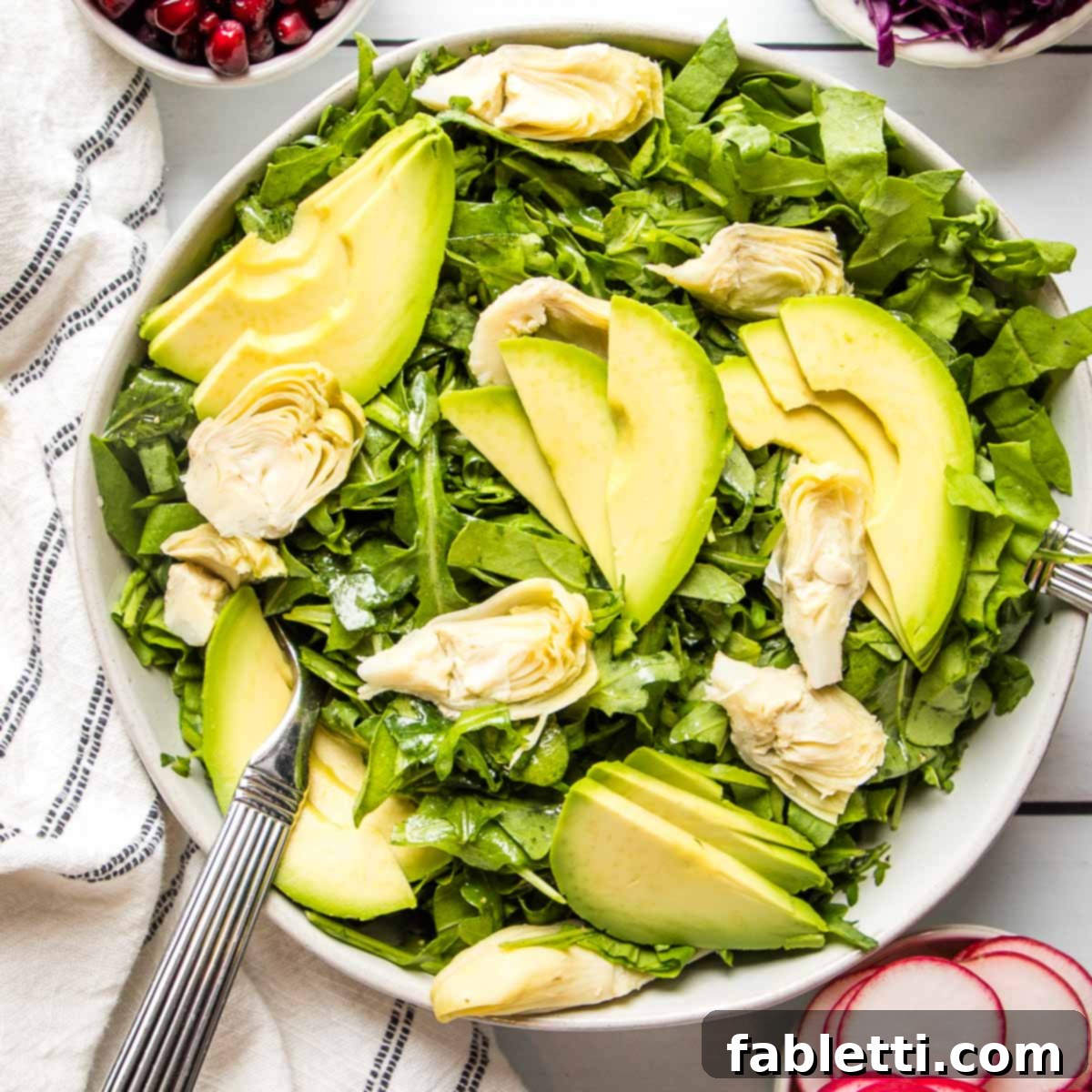 Vibrant Arugula & Lemon Salad 10 A large white bowl filled with dressed arugula, quartered artichoke hearts, and perfectly sliced avocado. Small separate bowls containing pomegranate seeds, sliced radishes, and chopped purple cabbage are arranged around the main salad, offering optional garnishes.