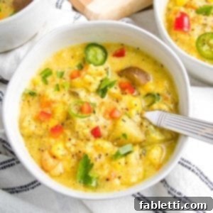 A beautiful white bowl, generously filled with creamy vegan corn chowder, adorned with colorful chunks of fresh vegetables. A spoon rests in the bowl, and the edges of other bowls of soup can be seen in the soft focus background. The chowder bowl sits atop a white linen napkin with elegant dark blue thin stripes.