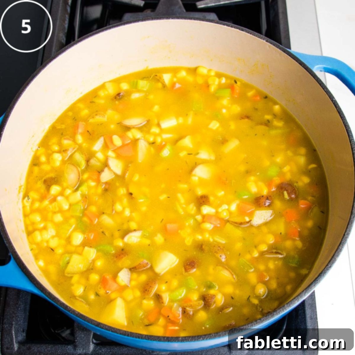 A vibrant yellow broth now covers the corn, potatoes, carrots, and celery in a blue Dutch oven, simmering gently.