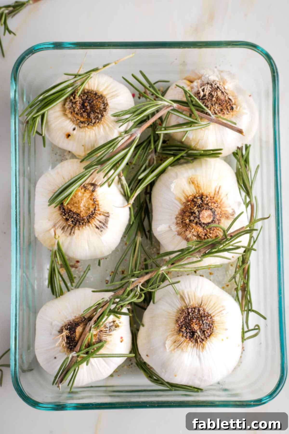 Sweet Creamy Oven Roasted Garlic 7 Six whole heads of garlic in a baking dish with fresh rosemary sprigs placed on top.