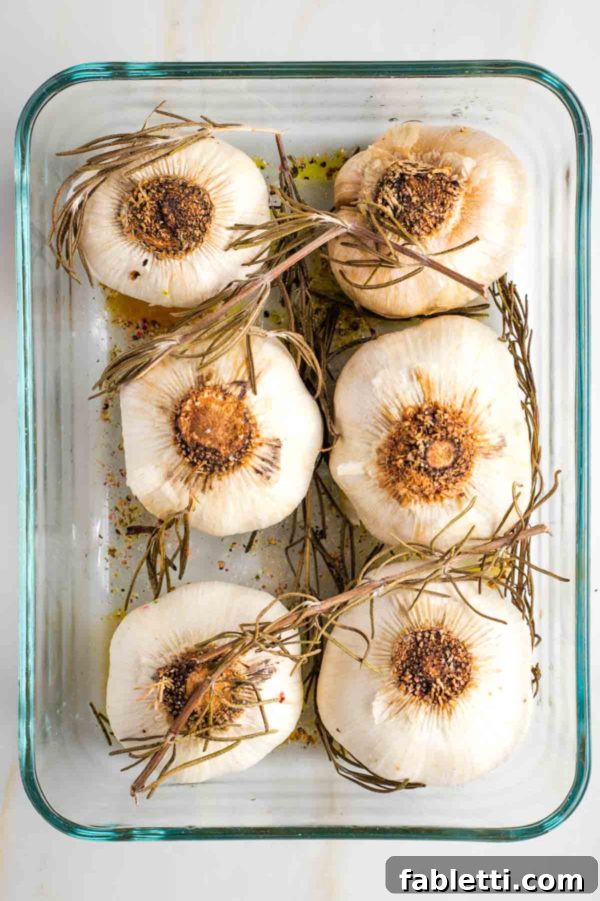 Sweet Creamy Oven Roasted Garlic 8 Garlic bulbs, now roasted to a golden hue, resting in a baking dish with wilted fresh rosemary.