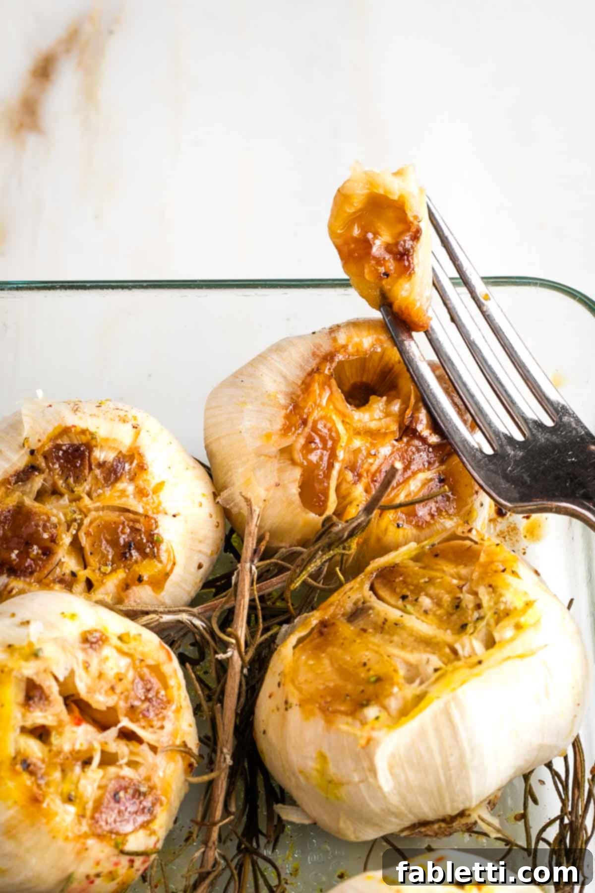 Sweet Creamy Oven Roasted Garlic 10 A fork pulling out a single, perfectly roasted clove of garlic from an entire bulb in a baking dish.