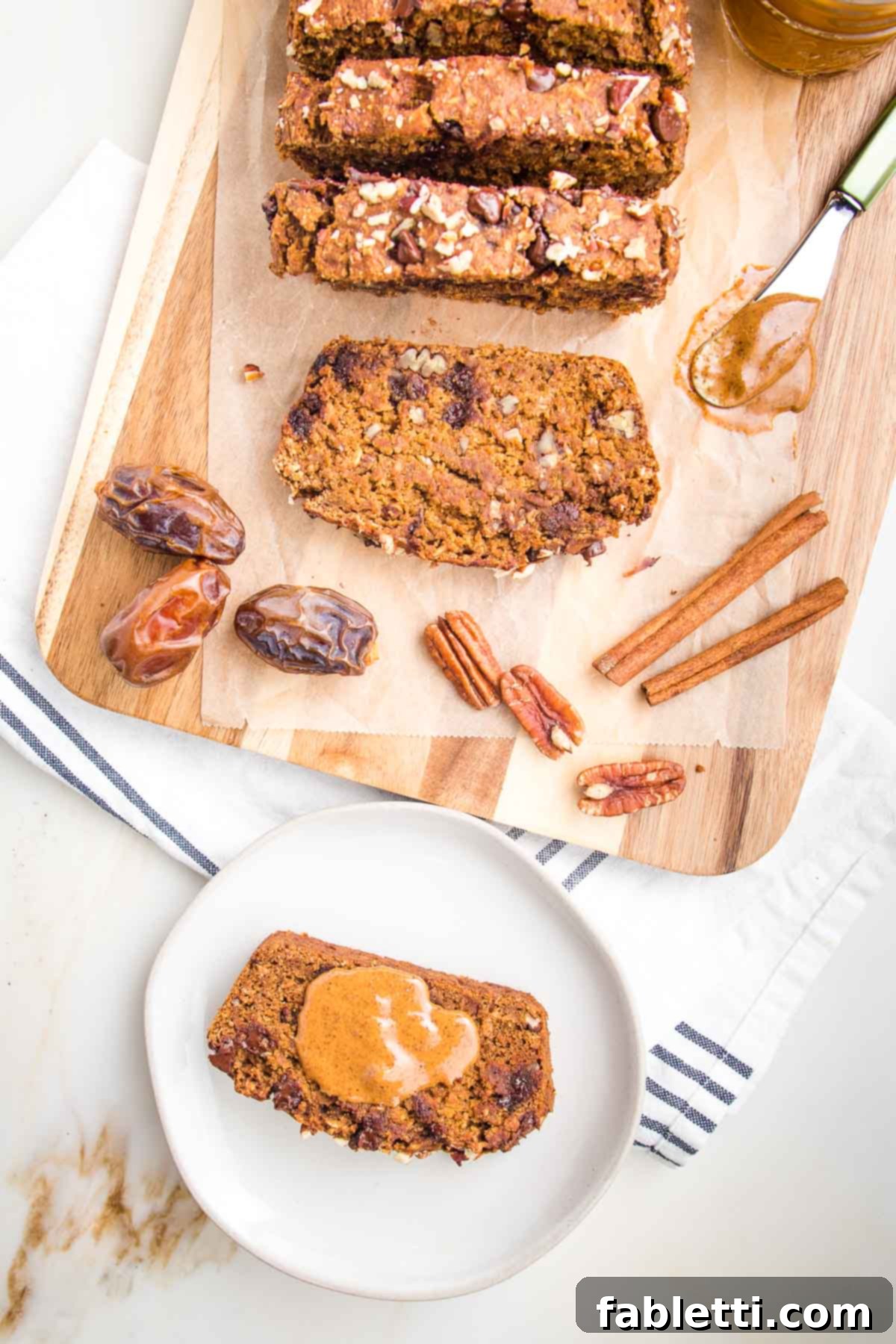 Harvest Spice Vegan Banana Loaf 2 Slices of fluffy vegan pumpkin bread with chocolate chips and pecans arranged on parchment paper on a cutting board. A single slice on a white plate with a dollop of almond butter sits in the foreground, ready to be enjoyed.