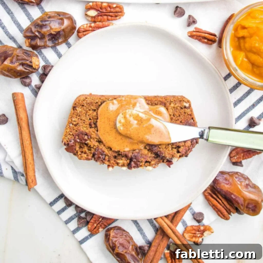 Harvest Spice Vegan Banana Loaf 11 A tantalizing slice of vegan pumpkin bread elegantly placed on a white plate, adorned with a dollop of creamy almond butter. The scene is beautifully accented with scattered Medjool dates, cinnamon sticks, pecans, and dark chocolate chips, evoking the cozy warmth of fall.