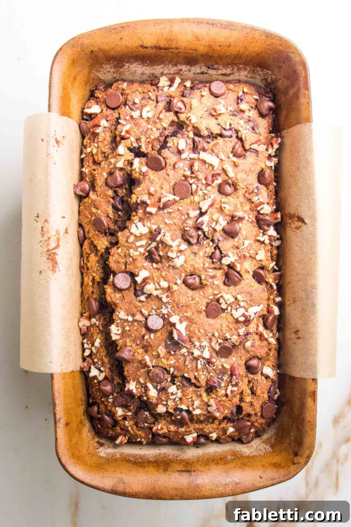 Harvest Spice Vegan Banana Loaf 8 A freshly baked pumpkin loaf, golden brown and still in its parchment-lined pan, just out of the oven, with melted chocolate chips and toasted pecans visible on top.