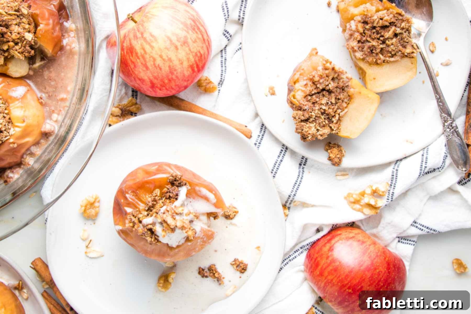 Naturally Sweet Dairy-Free Baked Apples 15 Edge of a glass pie dish filled with baked apples. A whole baked apple on a white rimmed plate with a small scoop of vanilla ice cream inside and another white plate with a whole baked apple cut in half so you can see the oatmeal cinnamon filling inside. Whole red apples, oat and walnut crumbs, and a navy striped tea towel are scattered around for an inviting presentation.
