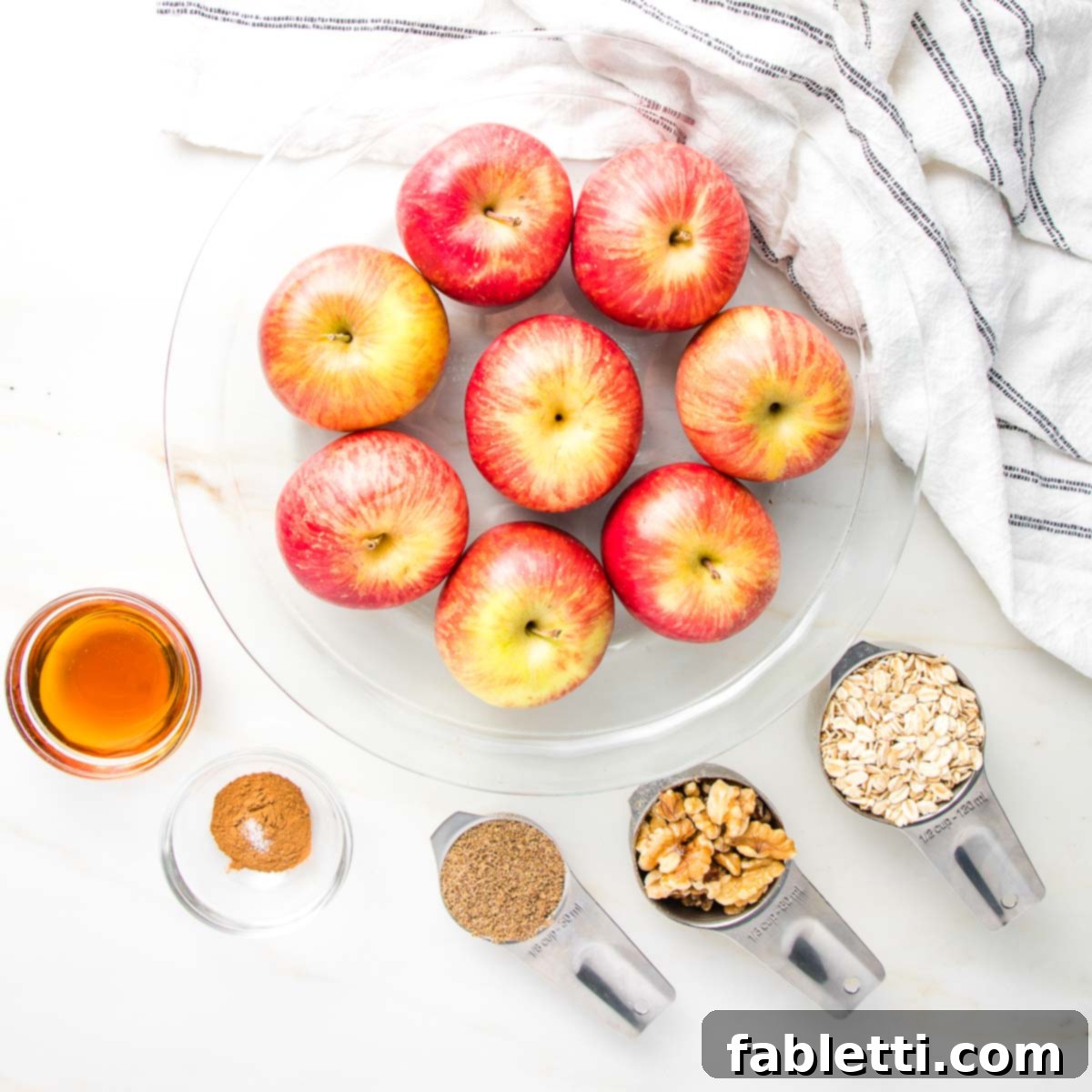 Naturally Sweet Dairy-Free Baked Apples 4 Glass pie plate filled with vibrant red apples. Ingredients for healthy baked apples: maple syrup, cinnamon, ground flax, walnuts, and rolled oatmeal, all laid out ready for preparation.