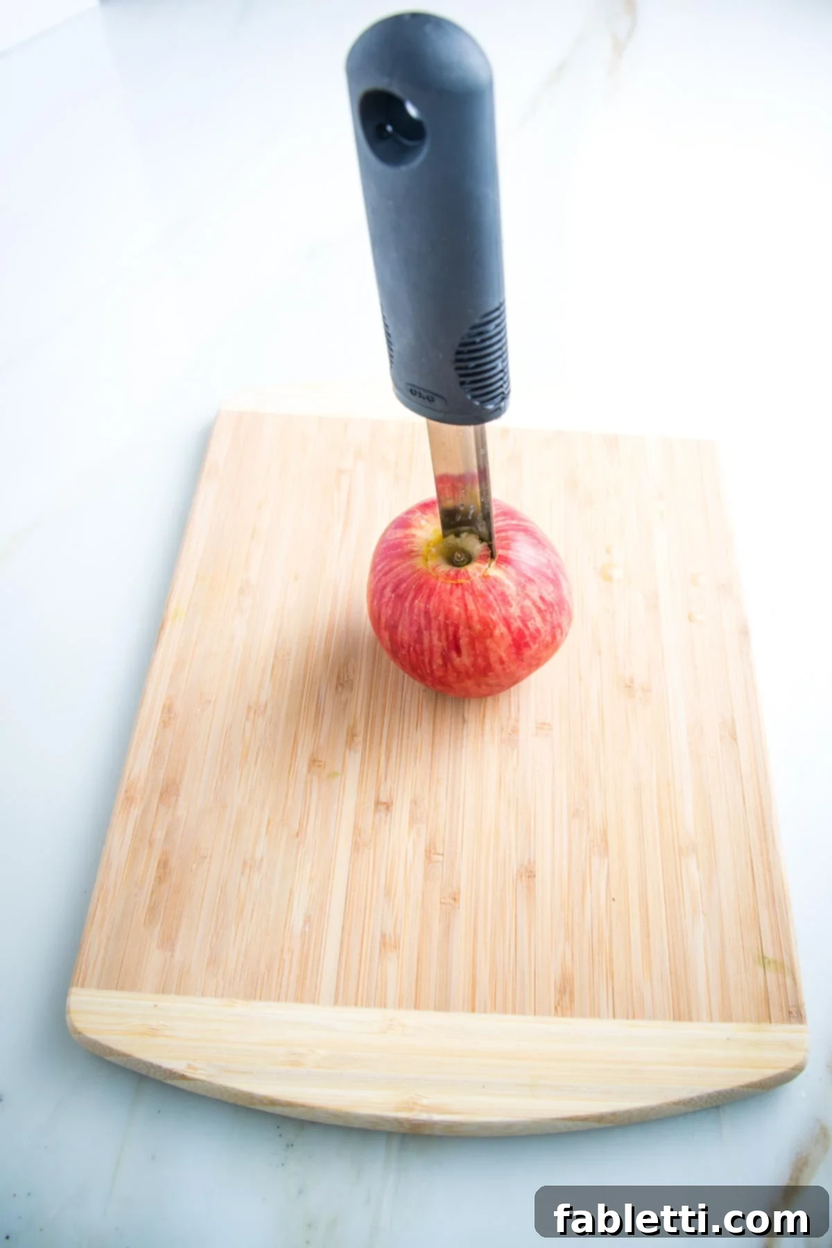Naturally Sweet Dairy-Free Baked Apples 5 Red apple on a cutting board with a metal apple corer inserted in the center, demonstrating the traditional method of preparing an apple for baking.