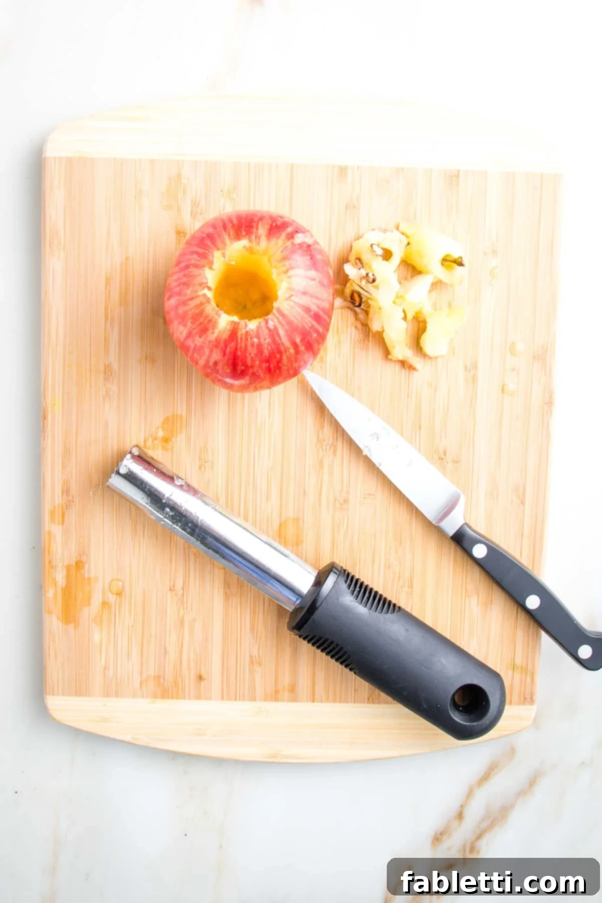 Naturally Sweet Dairy-Free Baked Apples 6 A cored red apple on a cutting board, with a paring knife and an apple corer lying beside it, showing the hollowed-out result of coring.