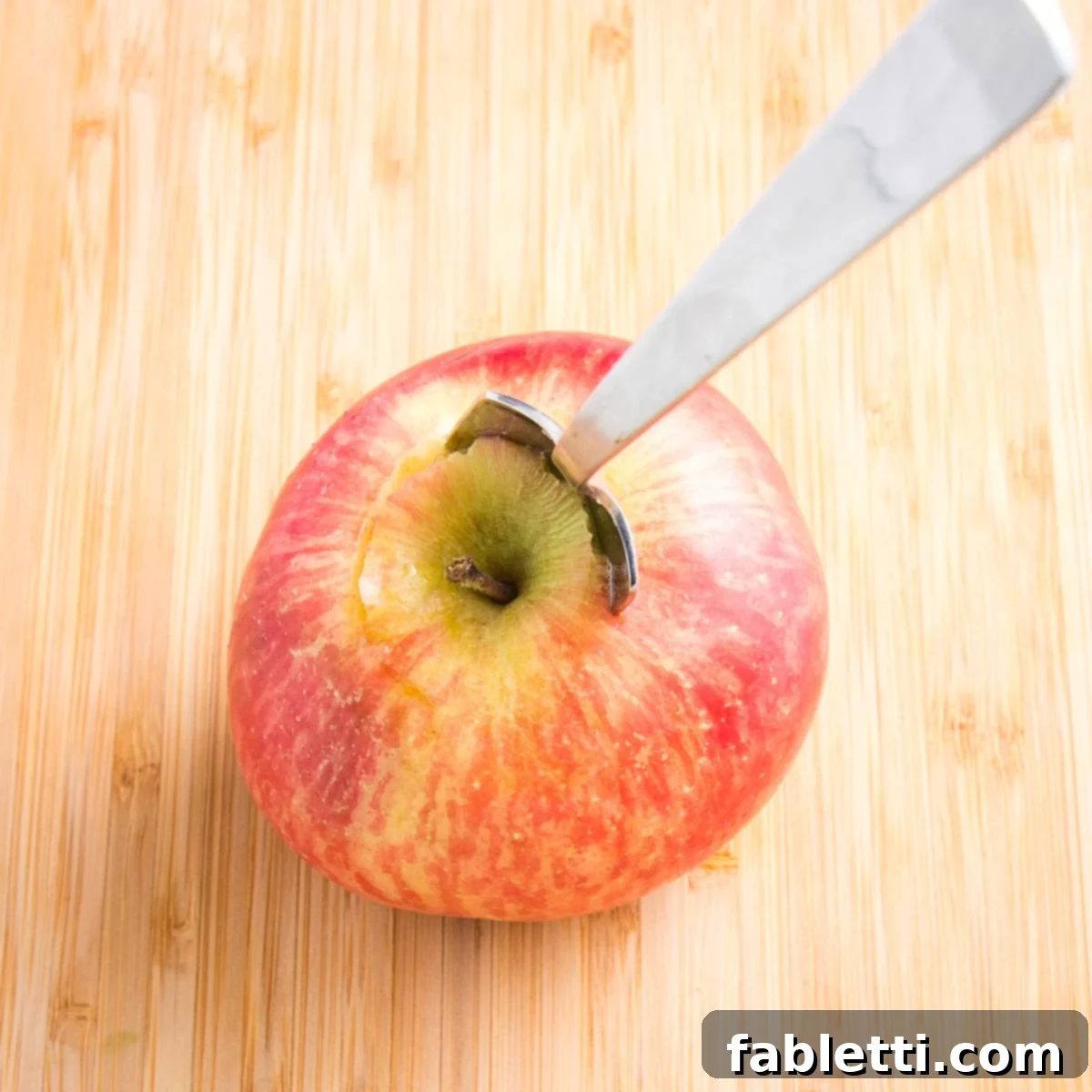 Naturally Sweet Dairy-Free Baked Apples 8 Close up of a red apple on a cutting board with a grapefruit spoon stuck into the center to scoop out the core, demonstrating an alternative, easier coring method.