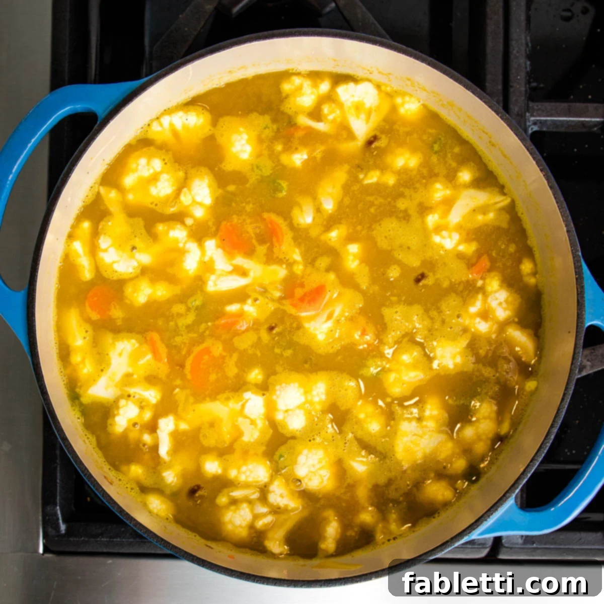 A vibrant blue Dutch oven is filled with steaming broth and an array of colorful vegetables, including tender cauliflower, simmering gently.