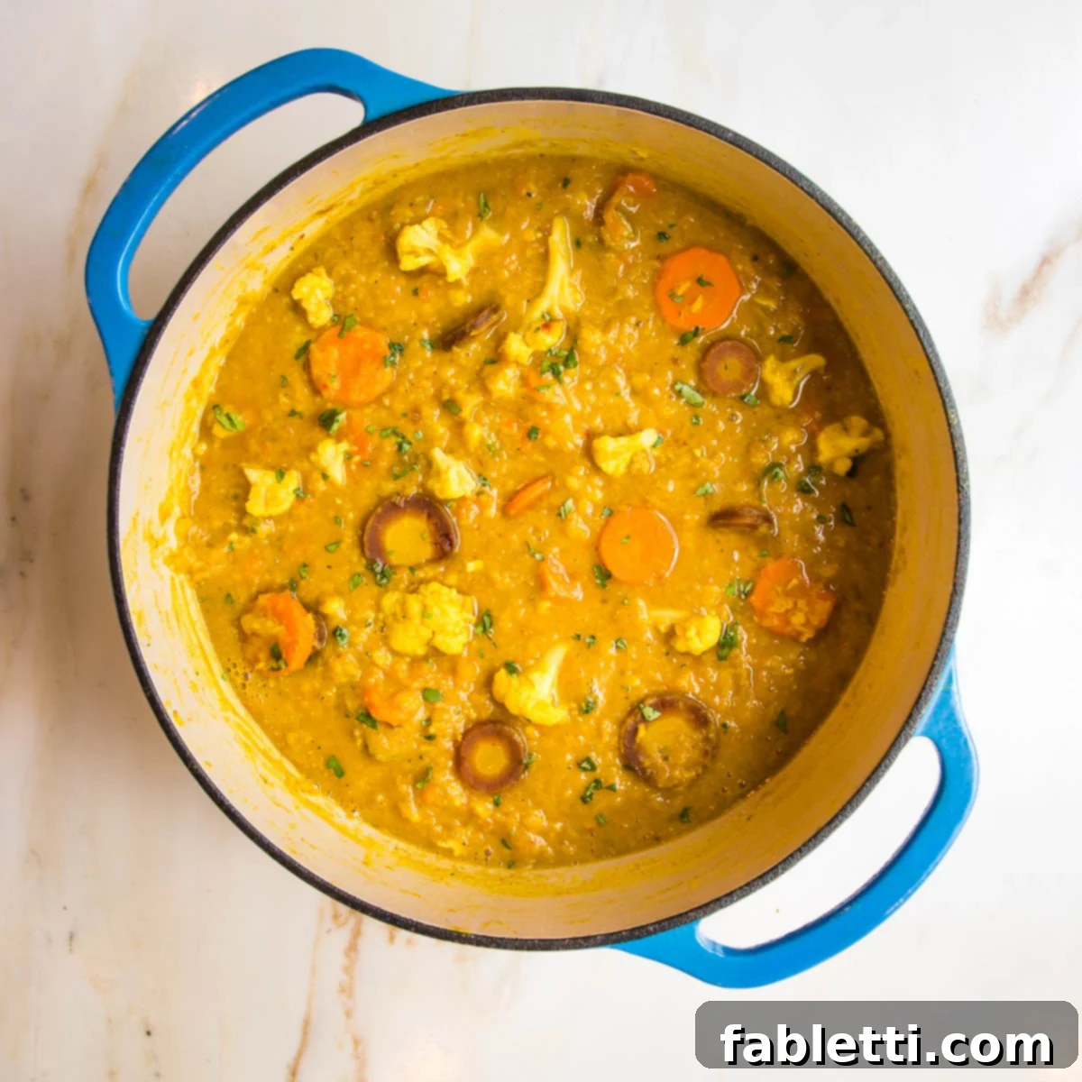 A blue Dutch oven proudly displays a generous serving of chunky yet creamy cauliflower red lentil soup, featuring enticing pieces of tender cauliflower and various colored carrots, promising a delightful texture.