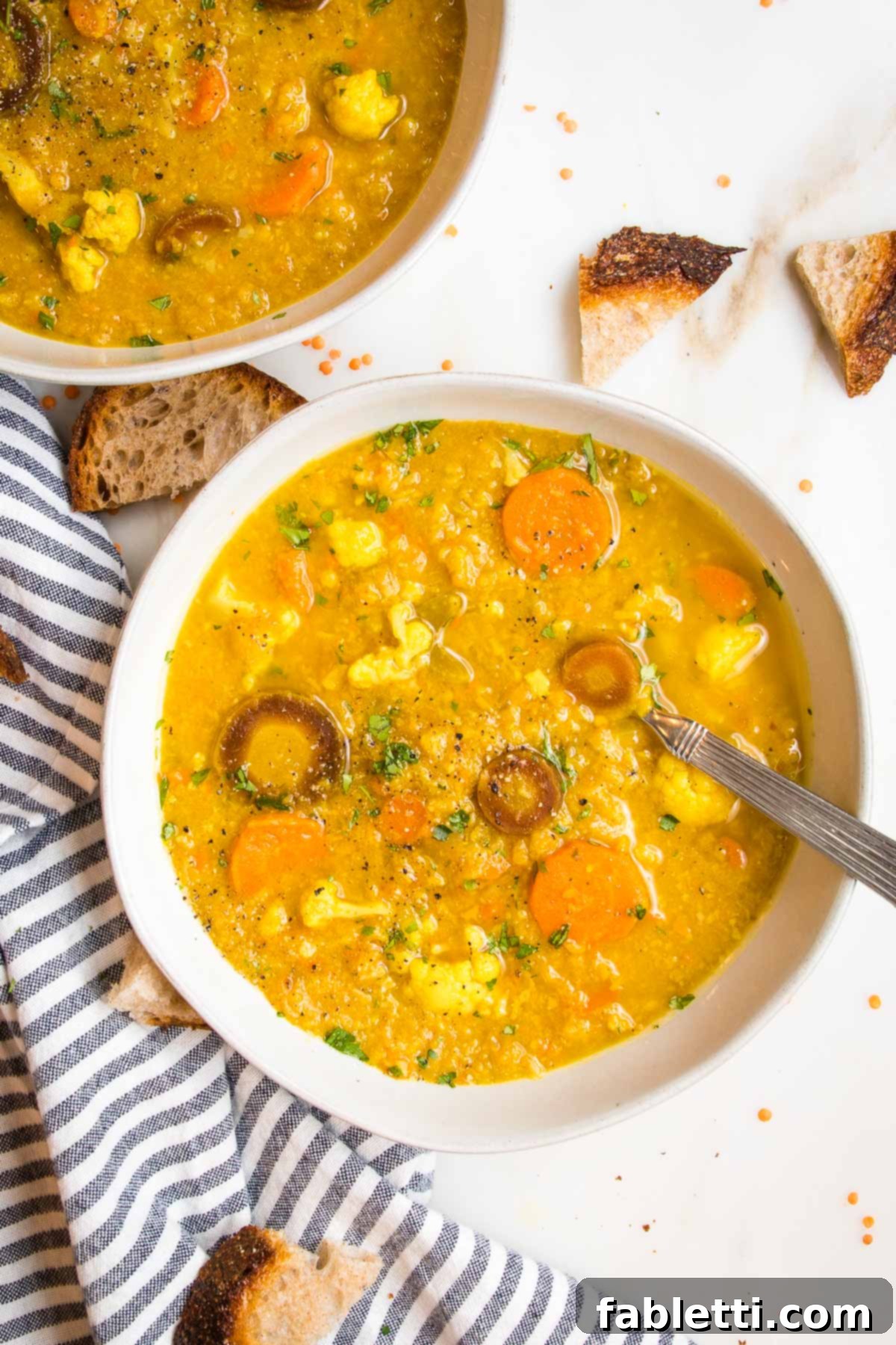 Large white bowl filled with yellow textured creamy soup with chunks of carrots and cauliflower. Crusty bread chunks on the table and a striped blue napkin.