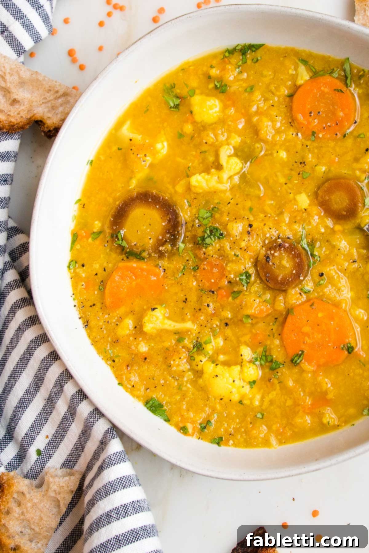 A beautifully presented white bowl of vibrant, colorful cauliflower red lentil soup. A charming blue striped cloth napkin rests gracefully to the side, and torn pieces of golden, crusty bread are artfully arranged along the edges, inviting you to dip.