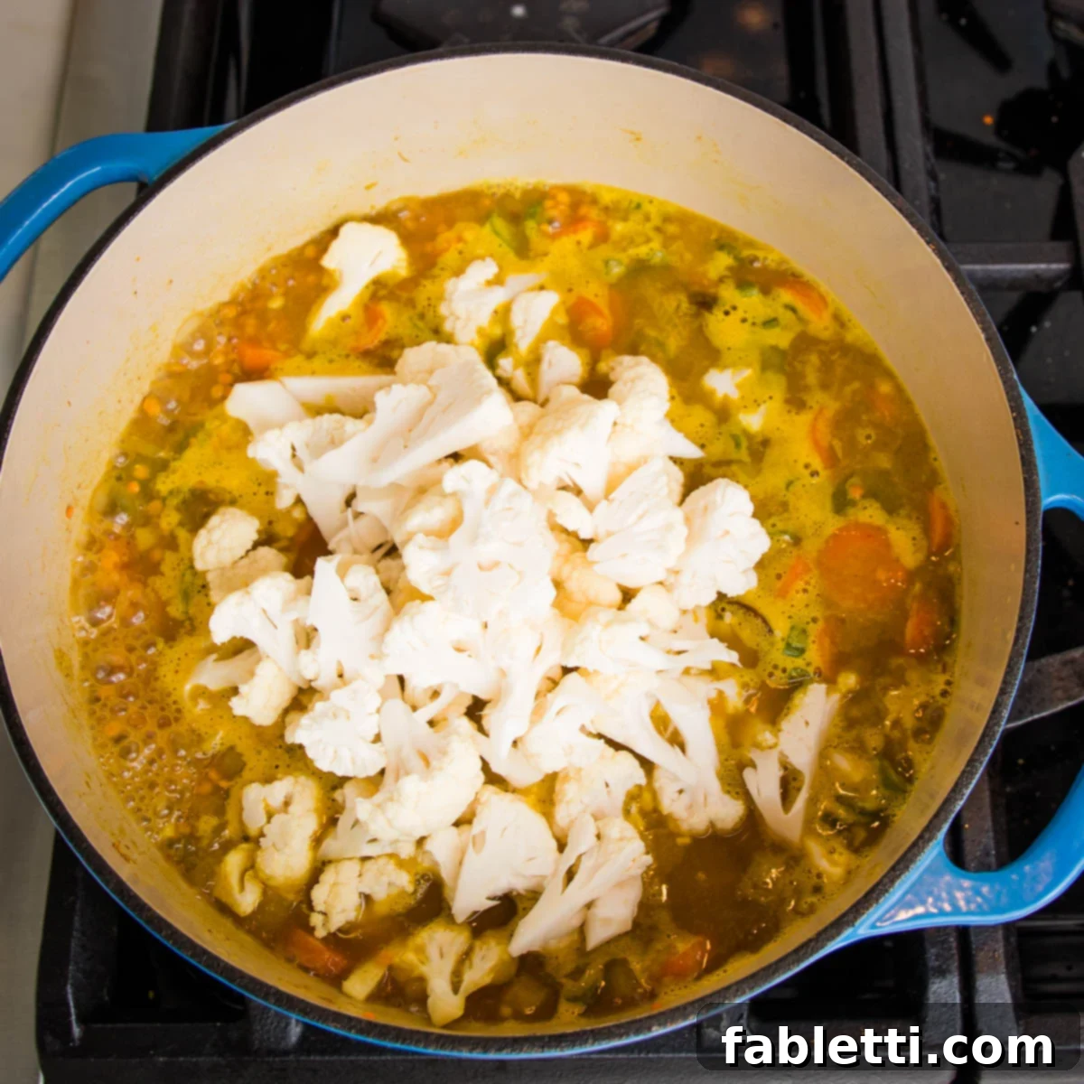 In the Dutch oven, cauliflower florets have just been added to the top of the cooking veggies and broth, ready to simmer and soften.