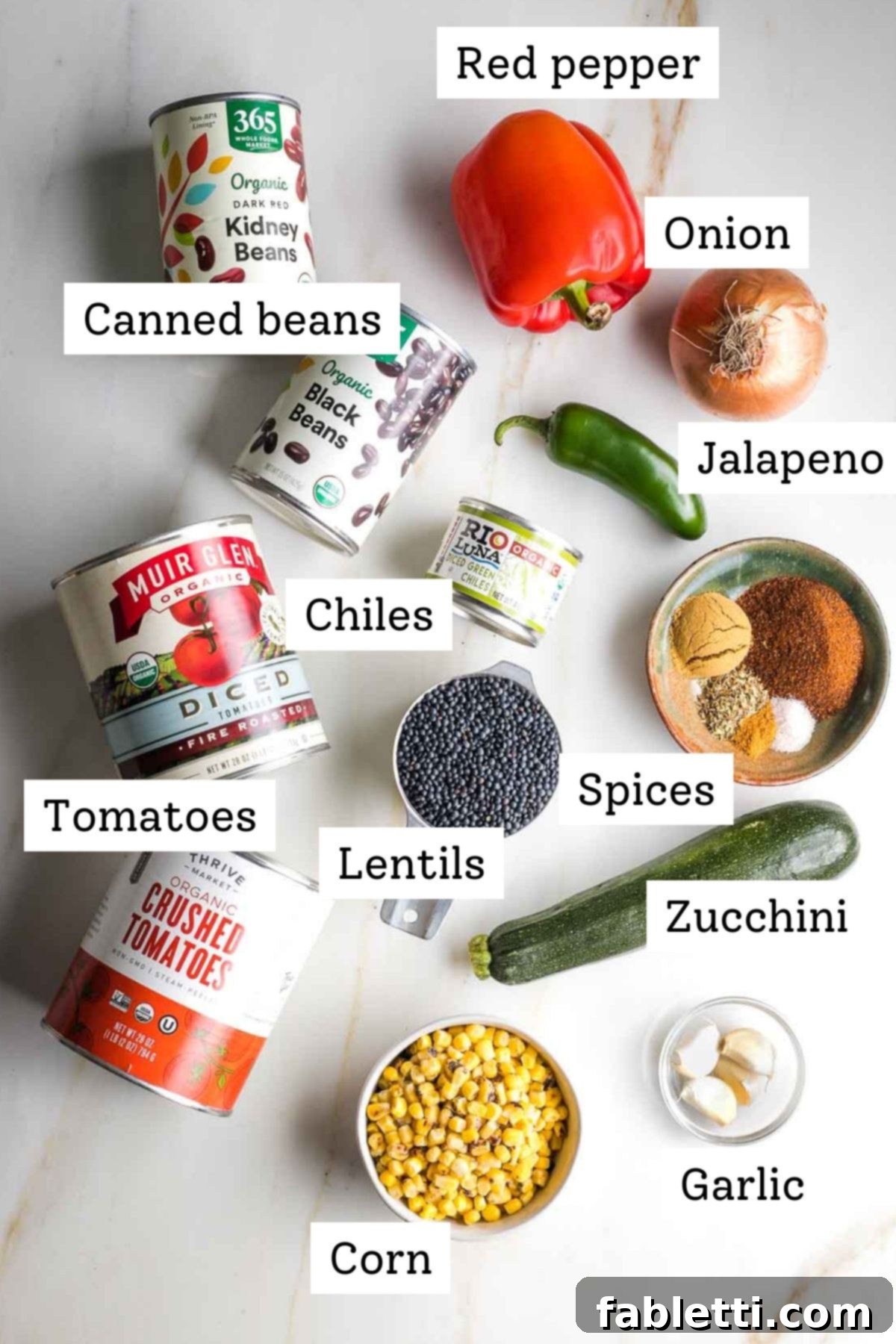 Quick and Flavorful Vegan Lentil Chili 4 A flat lay of fresh and canned ingredients for vegan chili: an assortment of canned beans, crushed and diced tomatoes, various spices in small bowls, a sliced yellow onion, red bell pepper, black beluga lentils, frozen corn kernels, minced garlic, and canned green chiles.