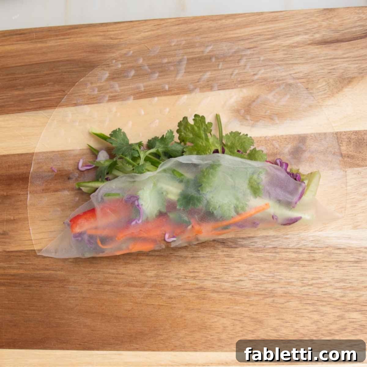 A softened rice paper wrapper filled with fresh vegetables, with the bottom edge gently lifted and folded upwards, initiating the rolling process.