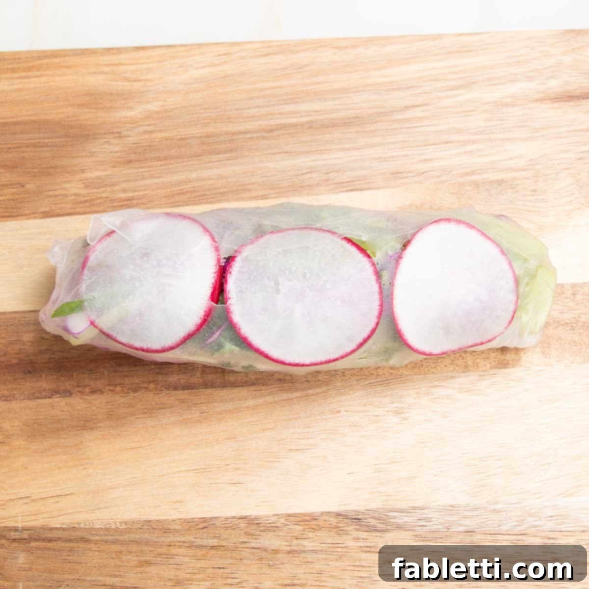 A perfectly rolled veggie spring roll featuring three thin slices of radish visible against its translucent wrapper, indicating a successful wrap.