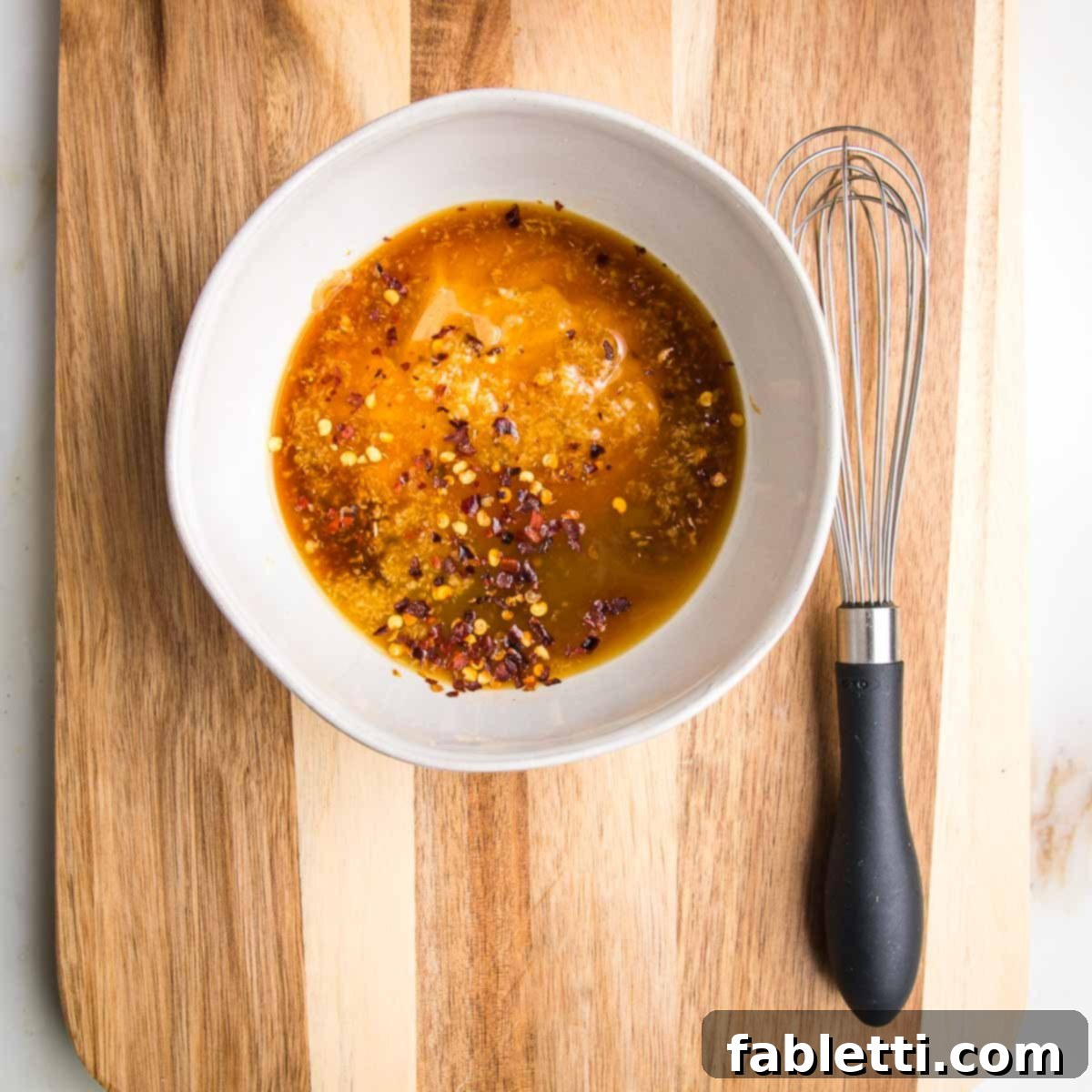 White bowl filled with peanut sauce ingredients: creamy peanut butter, tamari, rice vinegar, red pepper flakes, and freshly grated ginger. A balloon whisk rests beside the bowl.