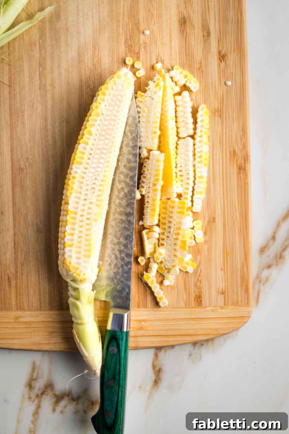 Zesty Israeli Corn and Pickle Salad 5 An ear of corn held horizontally on a cutting board, with a chef's knife carefully slicing off the kernels close to the cob.