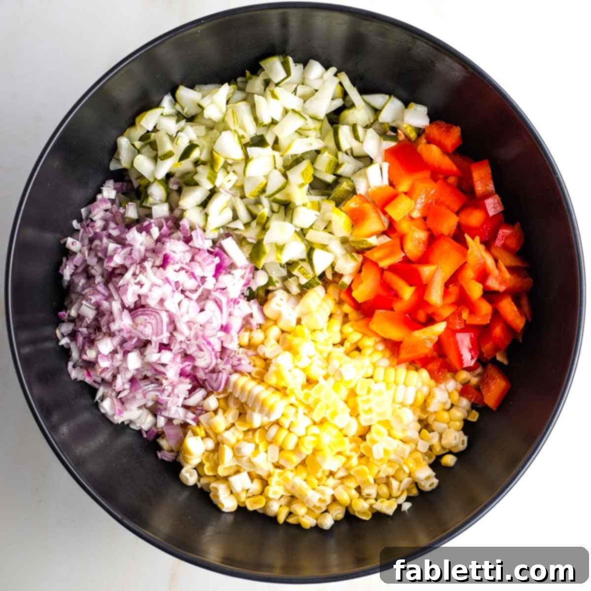 Zesty Israeli Corn and Pickle Salad 9 A large black mixing bowl containing colorful, distinct sections of diced pickles, shallots, corn kernels, and red bell peppers, ready to be combined.