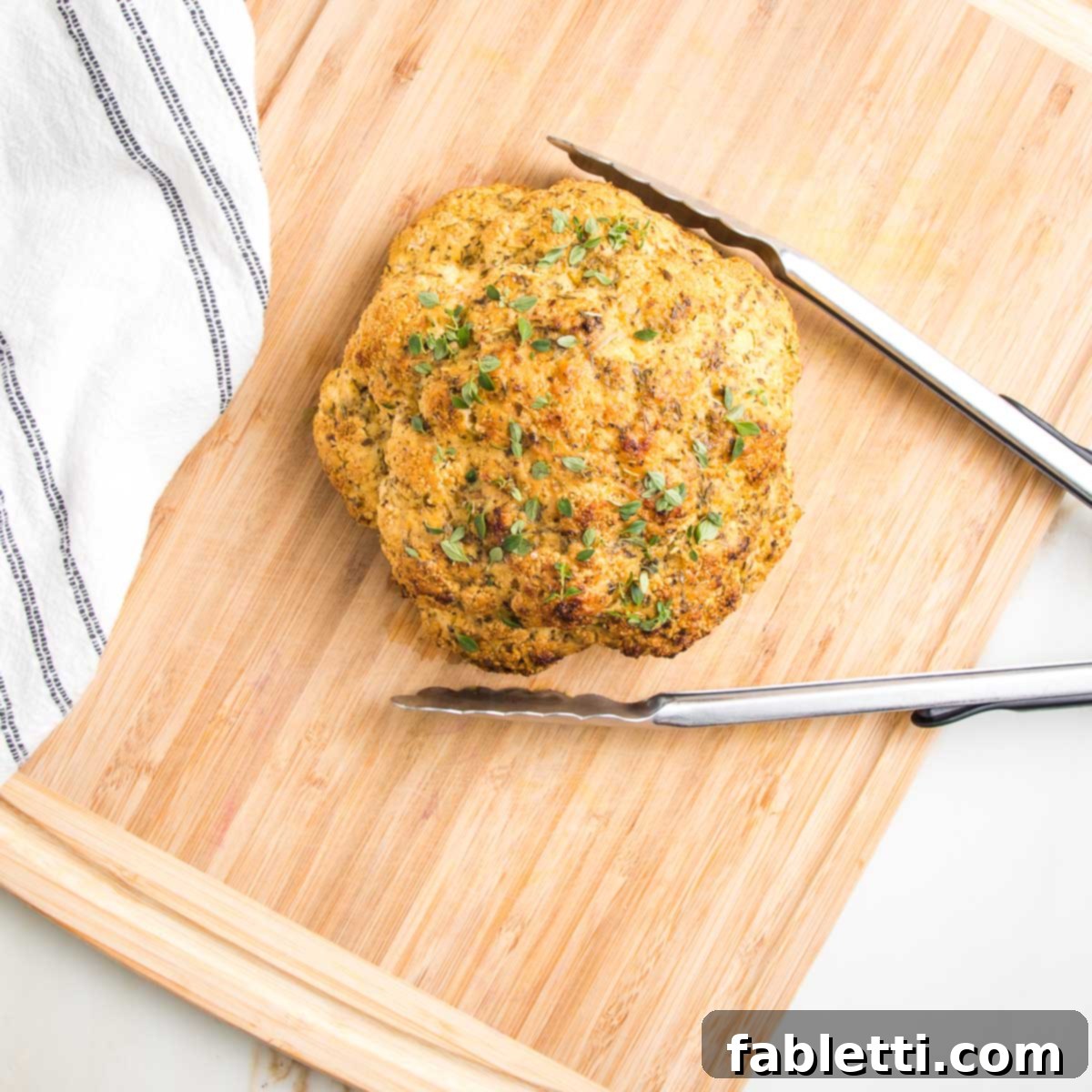 Whole roasted cauliflower with herbs on a cutting board with large tongs.
