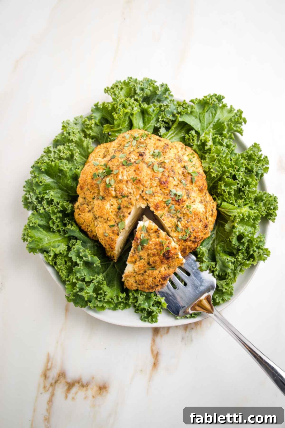 Whole roasted head of cauliflower on top of curly green kale on a white circular platter. A wedge has been cut and a spatula lifting out that piece.