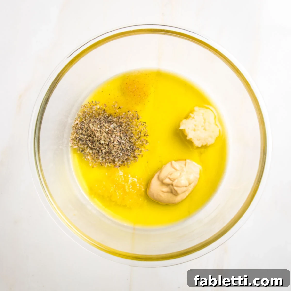 Medium sized glass bowl with olive oil and lemon juice and clumps of ingredients: dijon mustard, pressed garlic, italian spices, lemon zest, sea salt.