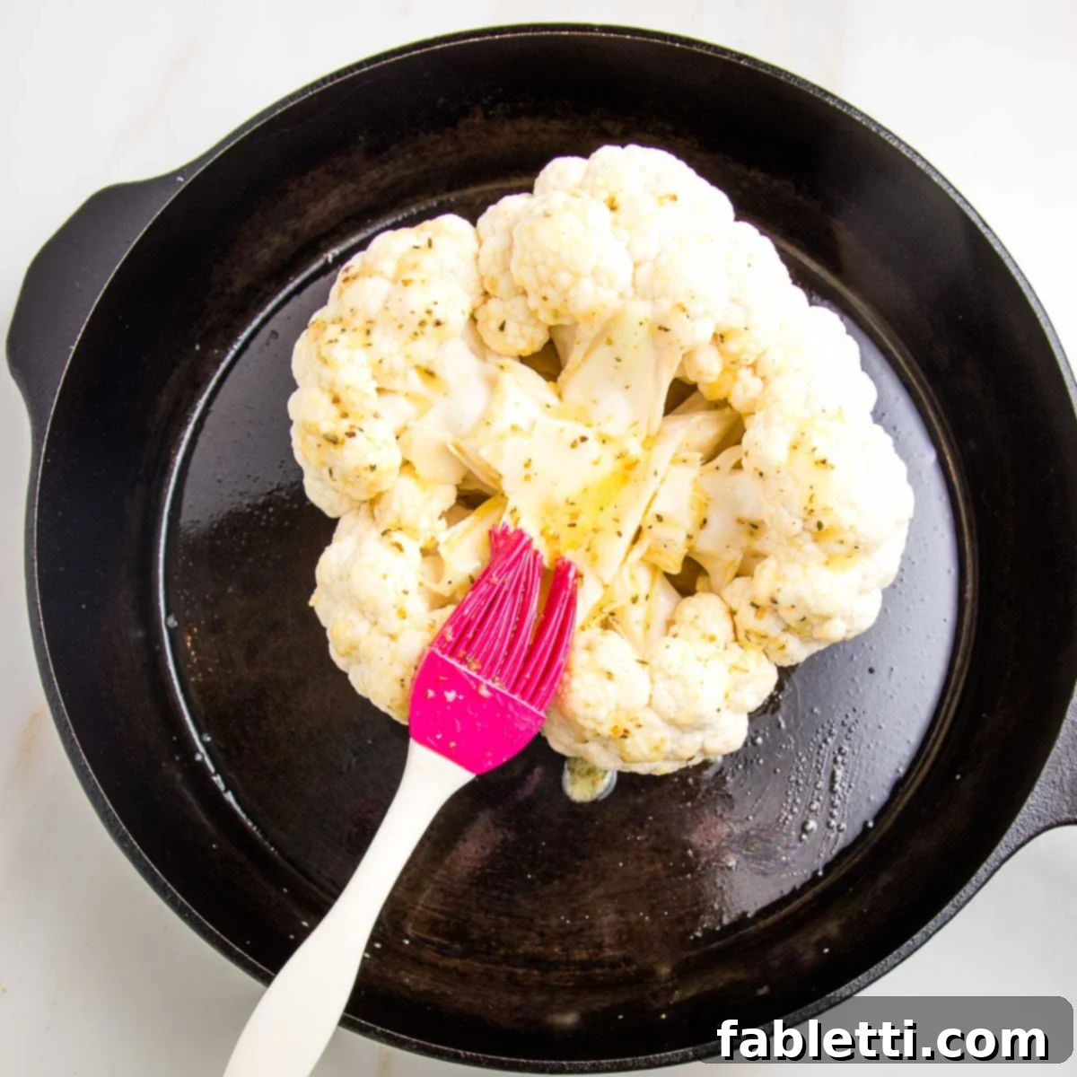 Cast Iron pan with whole head of cauliflower turned upside down, and a red silicone pastry brush basting on some marinade.