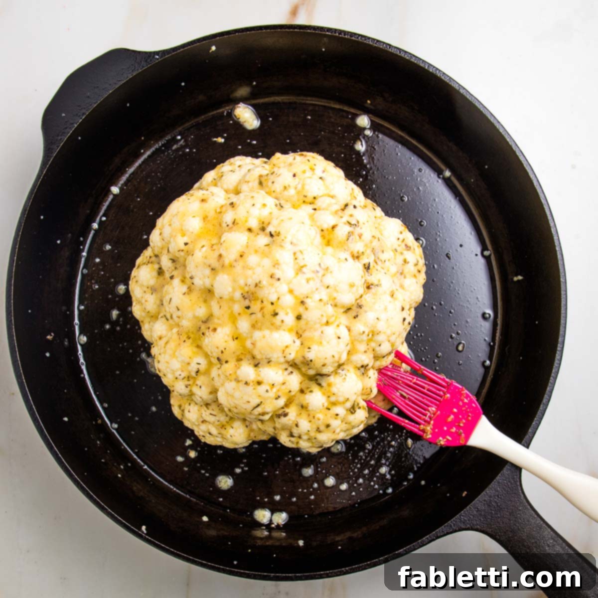 Cast iron pan with whole head of cauliflower and a red silicone pastry brush basting on lemon garlic herb marinade.