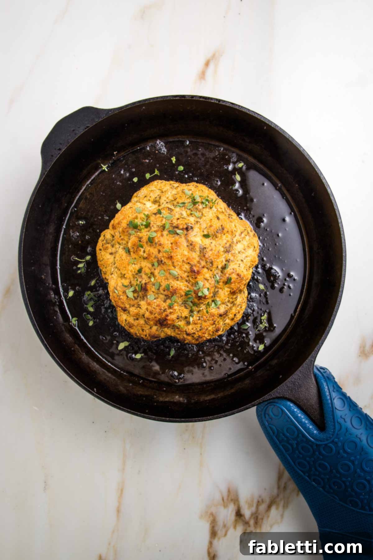 Cast iron skillet with a whole roasted head of cauliflower that's golden, and sprinkled with fresh thyme.