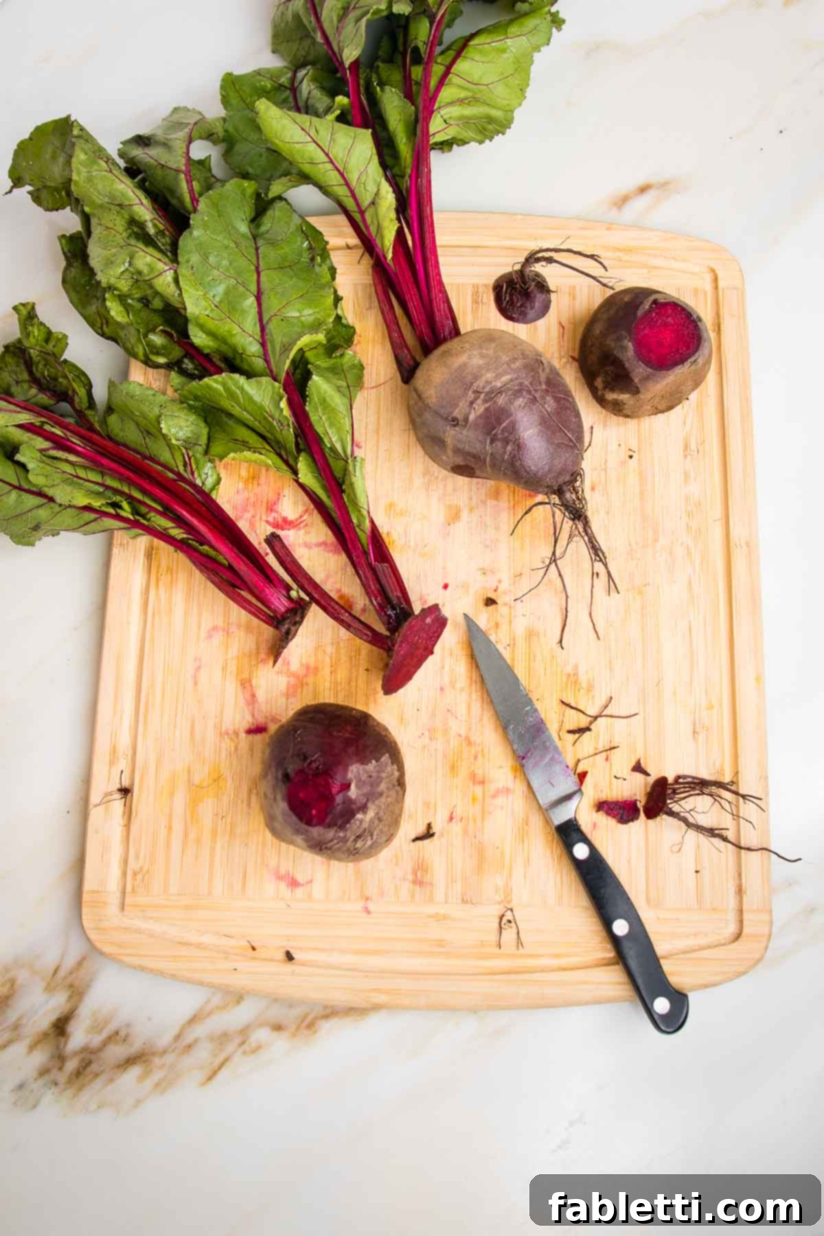 Balsamic Roasted Beet Salad 5 Trimming beet roots on a wooden board with a paring knife. You can see the beet greens and stems, plus three beets in various stages of trimming.