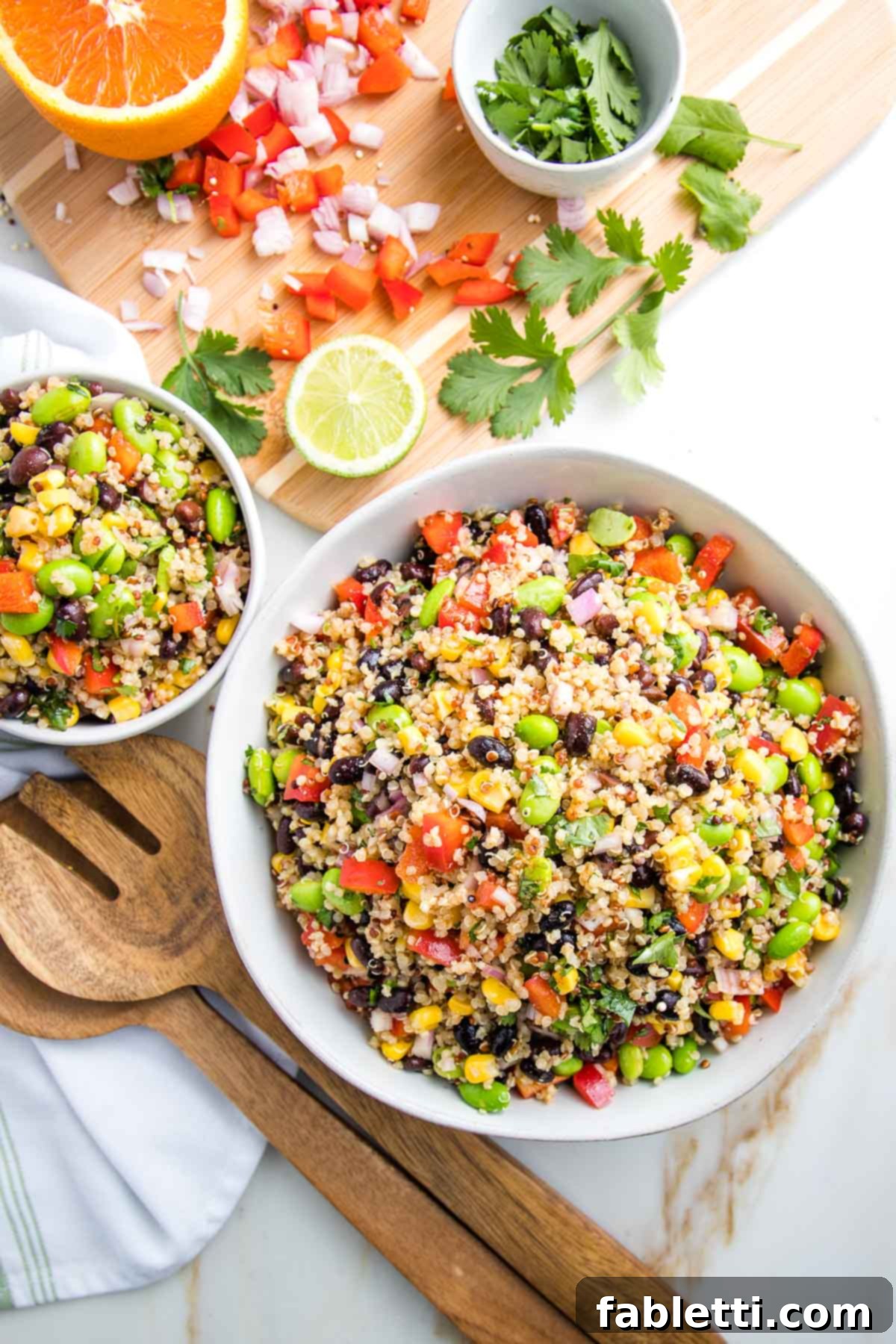 Zesty Southwest Quinoa Salad 2 Large and small white bowls filled with colorful quinoa salad. In the background there is a cutting board with scraps of chopped red pepper, shallot and fresh cilantro. Also there is half an orange and half a lime. Wooden salad servers are next to the bowls.