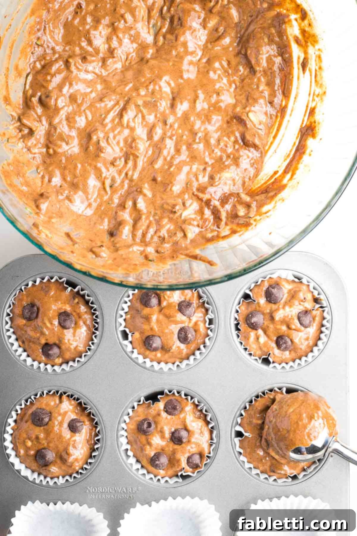 Decadent Double Chocolate Zucchini Muffins 10 Bowl of muffin batter being scooped into lined muffin tins and the muffins are topped with chocolate chips.