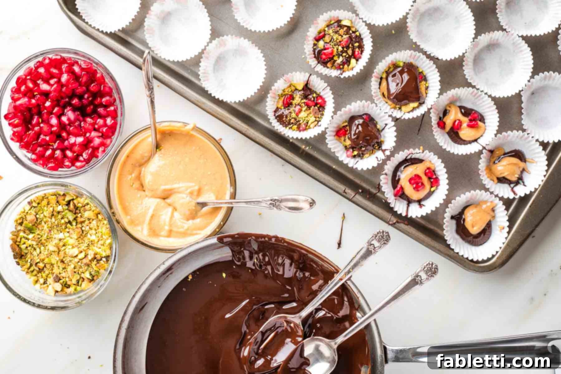 Jeweled Chocolate Peanut Butter Cups 8 Melted chocolate, chopped pistachios, peanut butter, and pomegranate seeds laid out near a baking sheet with assembled chocolate cups.