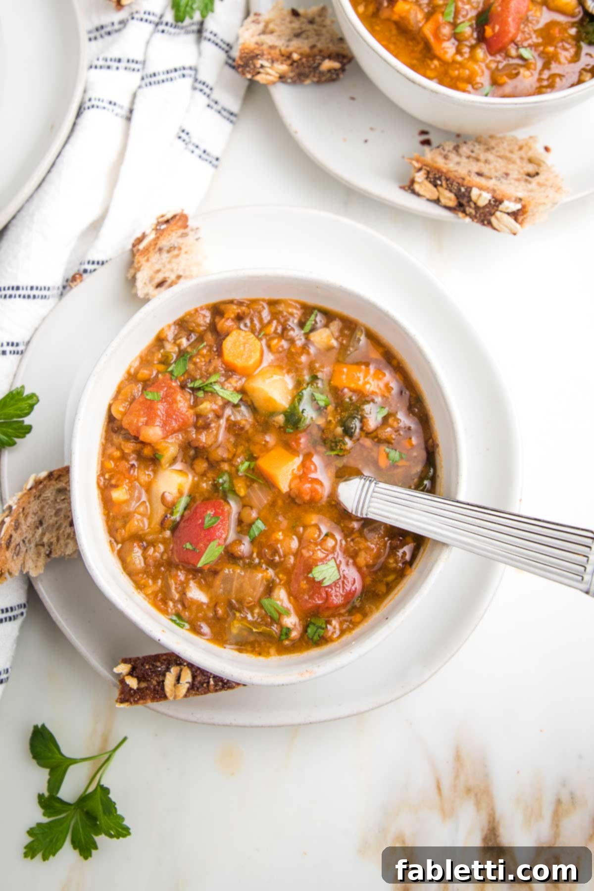Wholesome Plant-Based Lentil Soup 5 Bowl of chunky lentil soup, loaded with carrots, tomatoes, parsnips and celery in a white bowl. You can see part of another bowl in the distance. There are chunks of crusty bread and fresh parsley, plus a white dish towel with navy stripes.