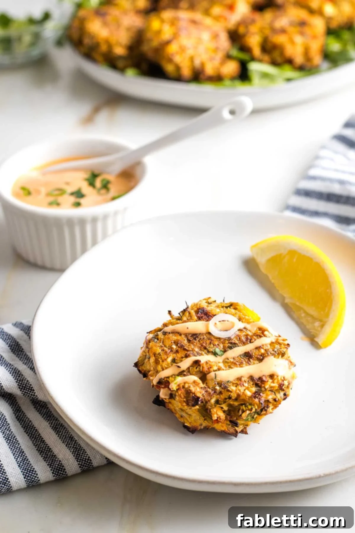 Vegan crab cake drizzled with sriracha sauce with a plateful of them in the background.