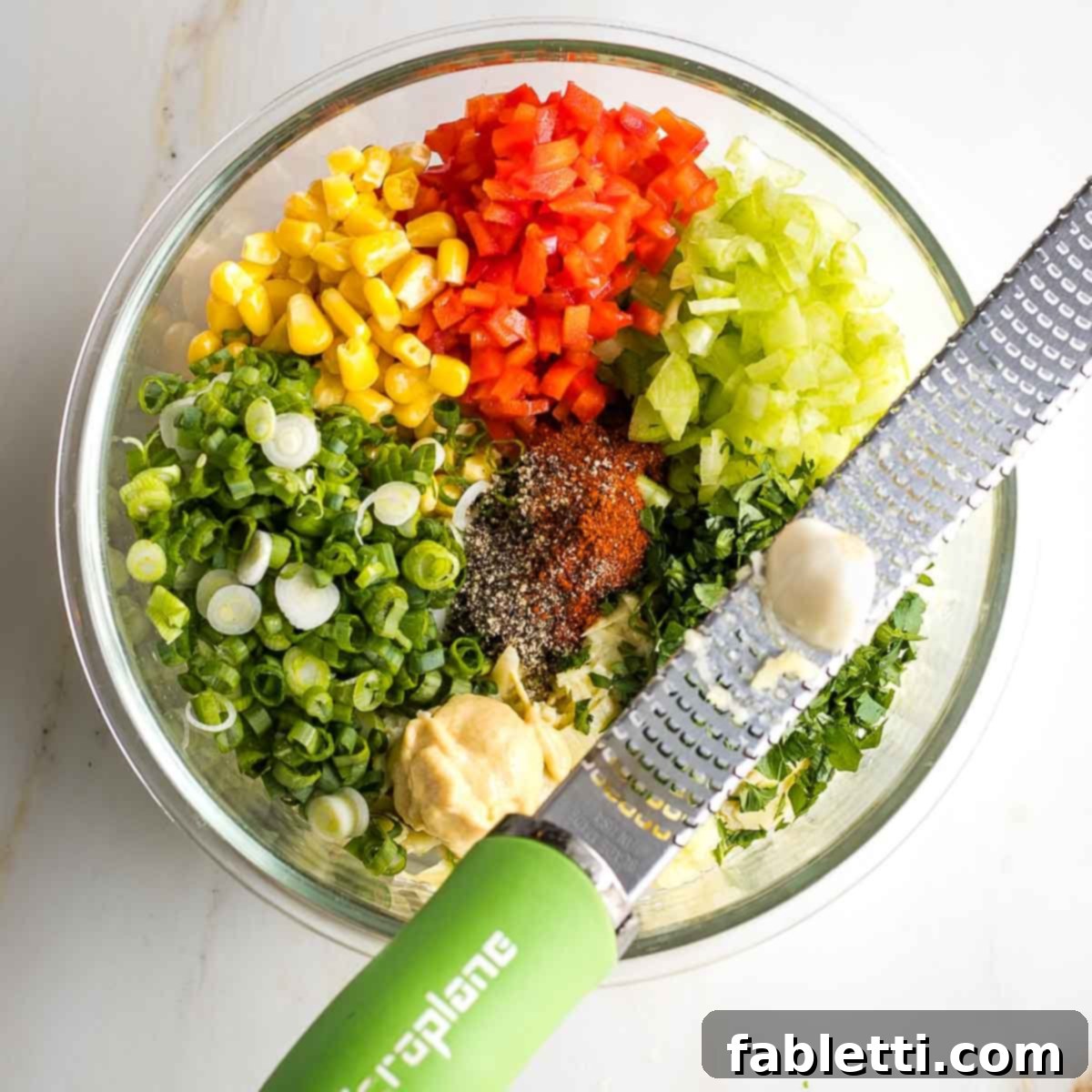 Grating garlic over bowl filled with red peppers, corn, scallions, celery, parsley, mustard and spices.