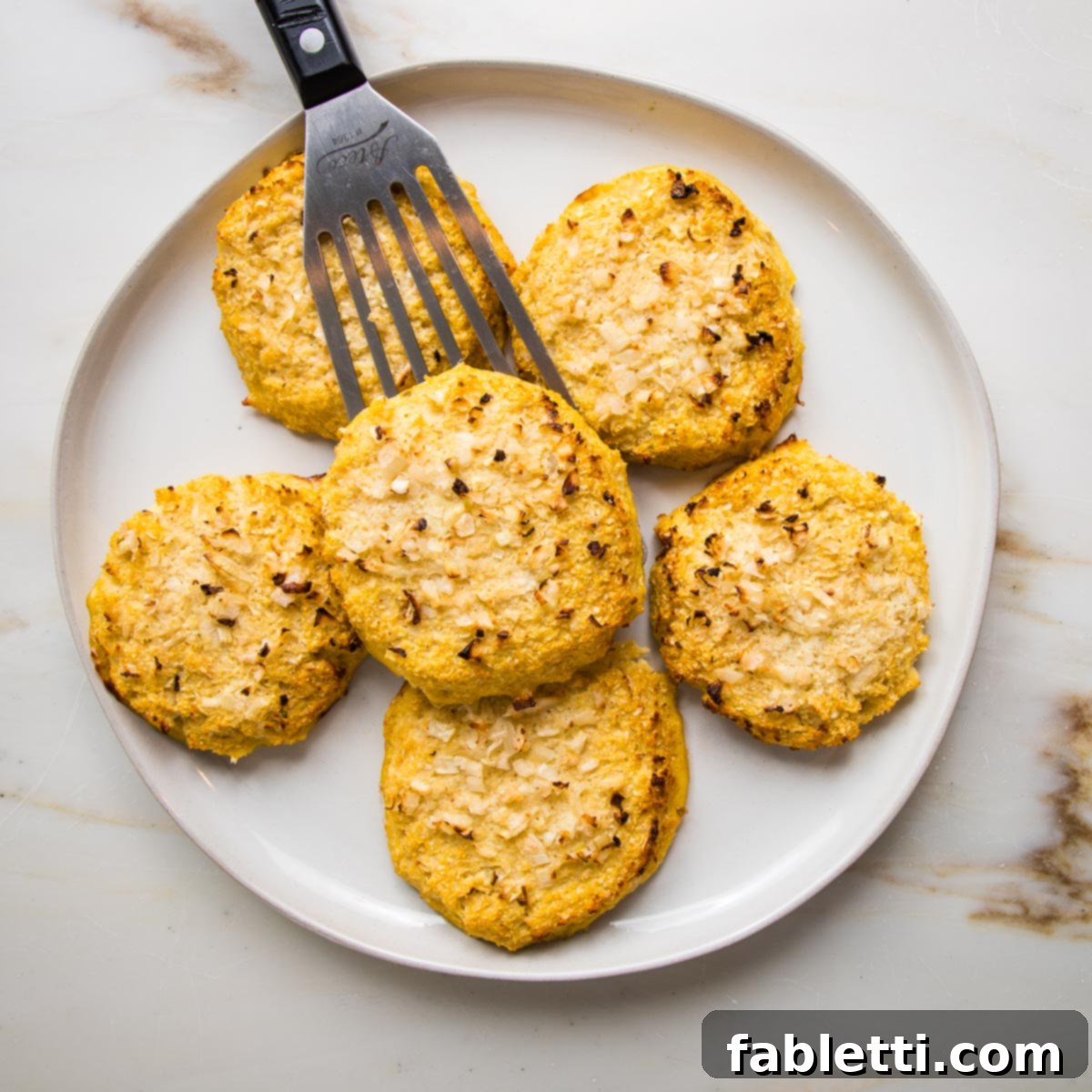 Flavorful Cauliflower Onion Buns 11 A white-rimmed plate is elegantly filled with several golden-brown cauliflower onion rolls, with a spatula poised to add one more, showcasing a delightful batch ready for serving.