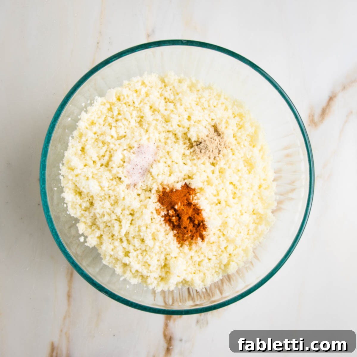 Flavorful Cauliflower Onion Buns 5 A glass bowl filled with finely riced cauliflower, lightly seasoned with spices sprinkled on top, ready for the next mixing step.